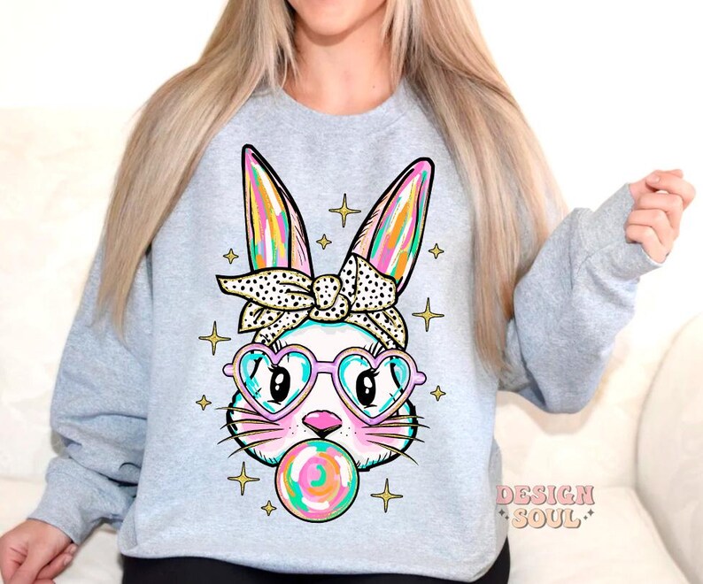 May include: A light gray sweatshirt with a colorful cartoon bunny wearing heart-shaped sunglasses and a bow. The bunny is holding a colorful lollipop. The text "DESIGN SOUL" is printed on the bottom right corner of the sweatshirt.