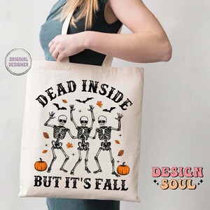 May include: A cream-colored tote bag with the text "DEAD INSIDE BUT IT'S FALL" in black. The design features three dancing skeletons, pumpkins, bats, and fall leaves. The bag is held by a person.