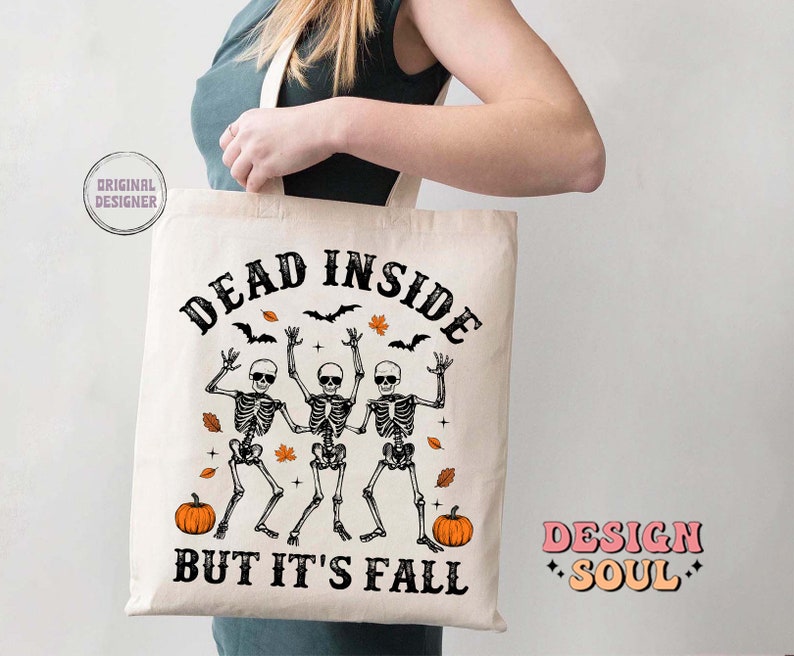 May include: A cream-colored tote bag with the text "DEAD INSIDE BUT IT'S FALL" in black. The design features three dancing skeletons, pumpkins, bats, and fall leaves. The bag is held by a person.