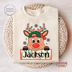 May include: A beige sweatshirt with a cartoon reindeer wearing a red and green hat. The reindeer is holding a sign that says "Jackson".