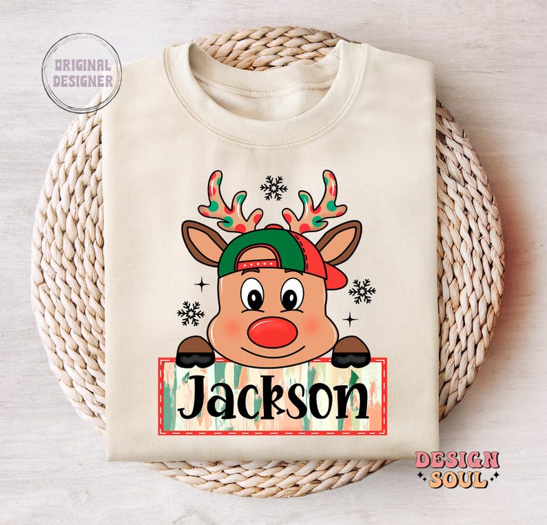 May include: A beige sweatshirt with a cartoon reindeer wearing a red and green hat. The reindeer is holding a sign that says "Jackson".
