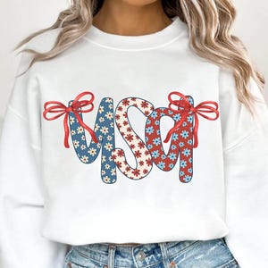 May include: White sweatshirt with a red, white, and blue floral pattern spelling out "USA" with red bows on either side.
