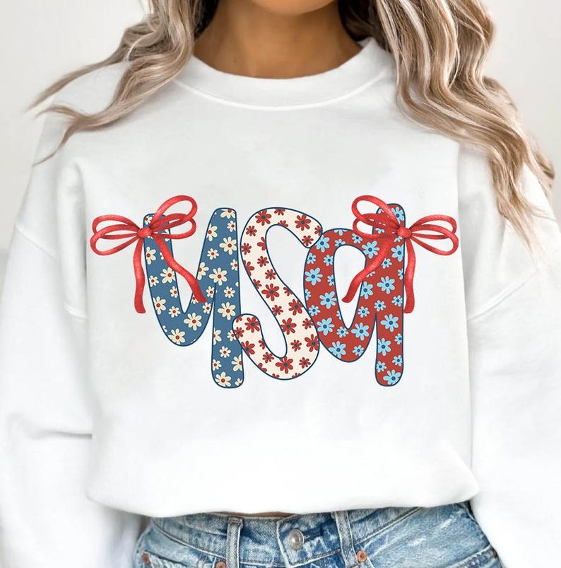 May include: White sweatshirt with a red, white, and blue floral pattern spelling out "USA" with red bows on either side.