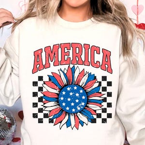 May include: White sweatshirt with a red, white, and blue sunflower graphic and the word "AMERICA" in red letters. The sunflower has a checkered background.