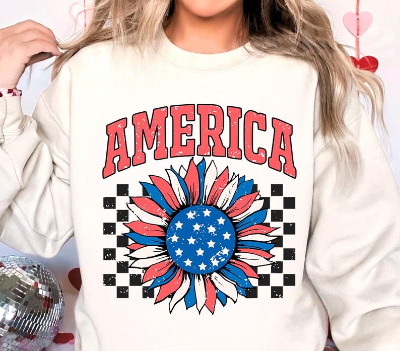 May include: White sweatshirt with a red, white, and blue sunflower graphic and the word "AMERICA" in red letters. The sunflower has a checkered background.