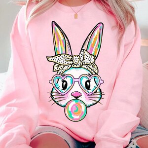 May include: Pink sweatshirt featuring a graphic of a white bunny wearing heart-shaped sunglasses and a polka dot headband. The bunny is holding a colourful lollipop.