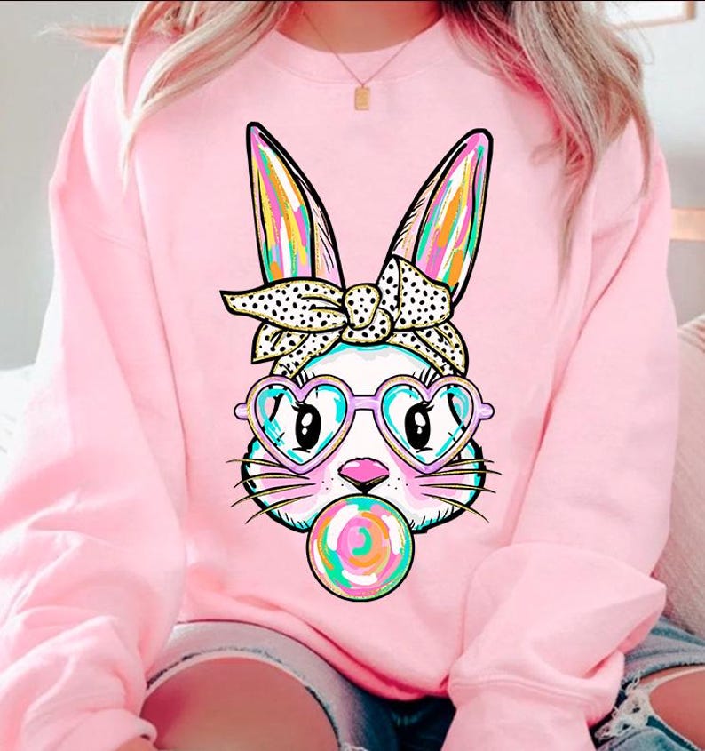 May include: Pink sweatshirt featuring a graphic of a white bunny wearing heart-shaped sunglasses and a polka dot headband. The bunny is holding a colourful lollipop.