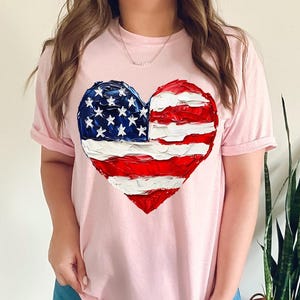 Pu&ograve; includere: Una maglietta rosa con una bandiera americana dipinta a forma di cuore.