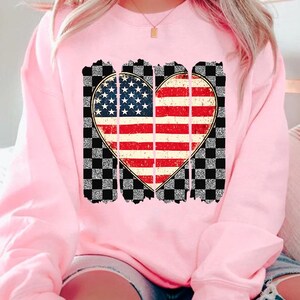May include: Pink sweatshirt with a distressed American flag heart design on a black and white checkered background.