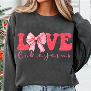 May include: A dark grey sweatshirt with the word "LOVE" in red featuring a pink bow in the centre and the words "like jesus" in white below.