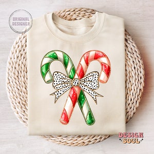 May include: A cream-colored sweatshirt with a design of two candy canes tied together with a black and white polka dot bow. The candy canes are red and white and green and white.