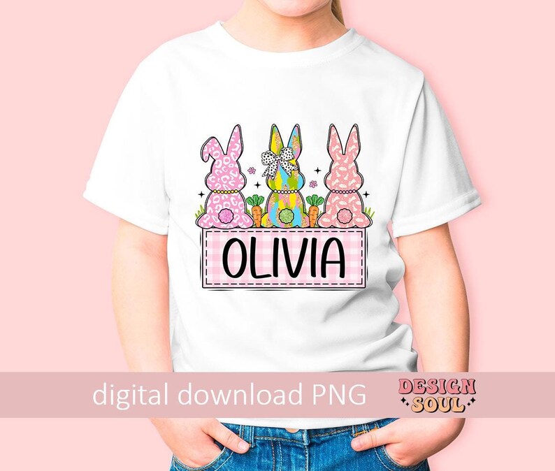 May include: White t-shirt with a pink, yellow, and blue bunny design. The bunnies are wearing bows and holding carrots. The text "Olivia" is on a pink and white checkered background.