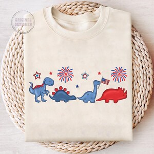 May include: Cream-colored sweatshirt featuring a row of cartoon dinosaurs in red and blue, with fireworks and stars. One dinosaur holds an American flag. The text "ORIGINAL DESIGNER" is in the upper left corner.