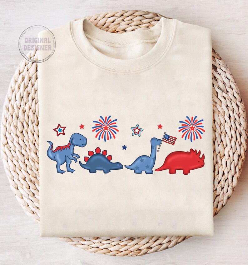 May include: Cream-colored sweatshirt featuring a row of cartoon dinosaurs in red and blue, with fireworks and stars. One dinosaur holds an American flag. The text "ORIGINAL DESIGNER" is in the upper left corner.