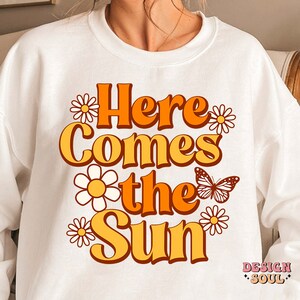 May include: White sweatshirt with a retro-style graphic design that says "Here Comes the Sun" in orange and yellow letters with white daisies and a butterfly.