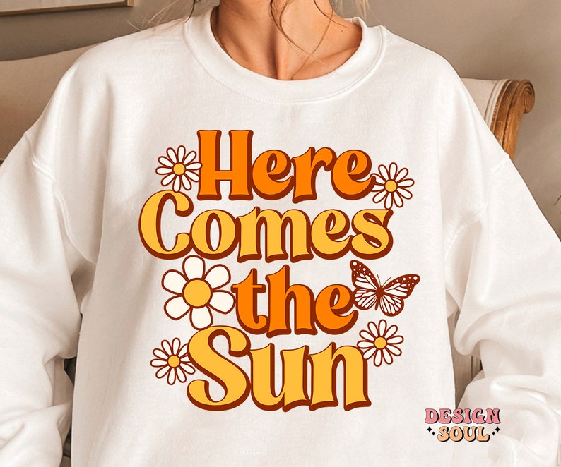 May include: White sweatshirt with a retro-style graphic design that says "Here Comes the Sun" in orange and yellow letters with white daisies and a butterfly.