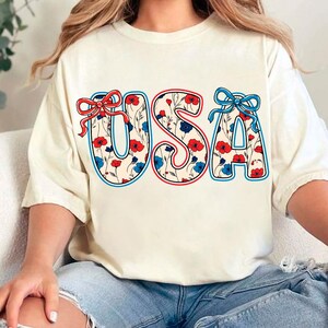 May include: A white t-shirt with a floral pattern design that spells out "USA" in red, white, and blue. The letters are outlined in red and blue.