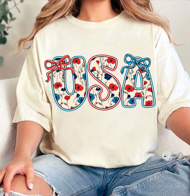 May include: A white t-shirt with a floral pattern design that spells out "USA" in red, white, and blue. The letters are outlined in red and blue.