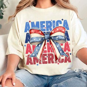 May include: Cream-colored t-shirt with the word "AMERICA" in blue and red block letters. A large bow in red, white, and blue with stars is centered on the shirt. The shirt is casual and suitable for patriotic events.