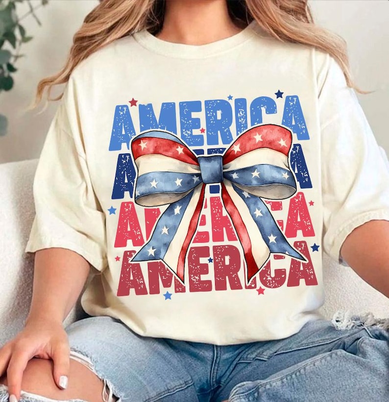 May include: Cream-colored t-shirt with the word "AMERICA" in blue and red block letters. A large bow in red, white, and blue with stars is centered on the shirt. The shirt is casual and suitable for patriotic events.
