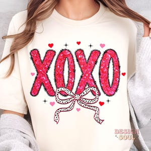 May include: A white t-shirt with a pink glitter XOXO design and a black and white polka dot bow. The shirt has a crew neck and short sleeves.