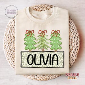 May include: A cream-colored sweatshirt with a Christmas tree design. The trees are green with red bows and the name "Olivia" is printed in a leopard print pattern.