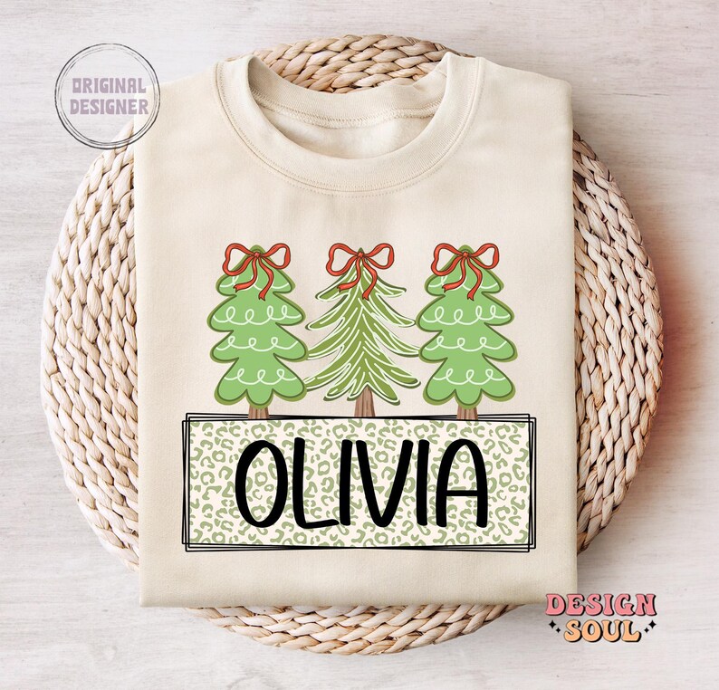 May include: A cream-colored sweatshirt with a Christmas tree design. The trees are green with red bows and the name "Olivia" is printed in a leopard print pattern.