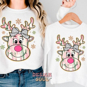 May include: White long-sleeved shirts with a pink and green reindeer design. The reindeer has a pink nose, black eyes, and a white and black bow. The design is surrounded by gold snowflakes.