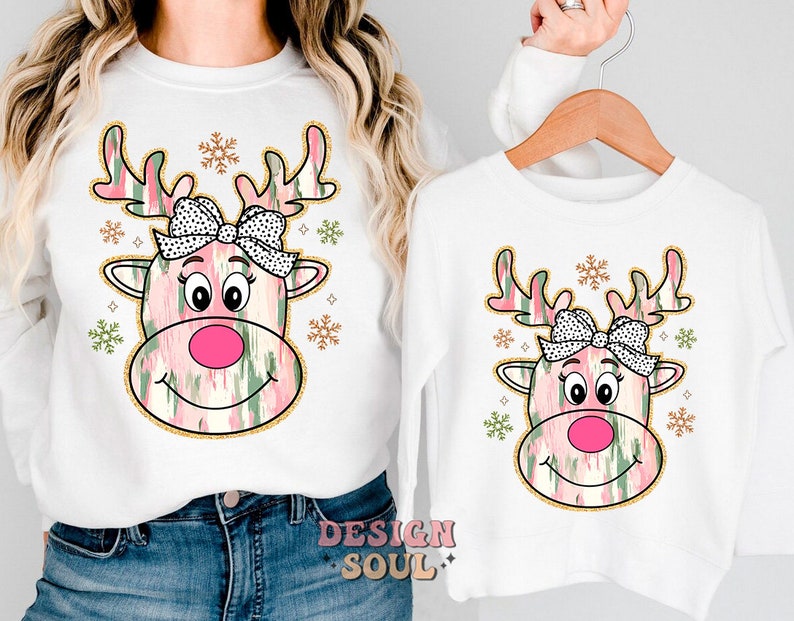 May include: White long-sleeved shirts with a pink and green reindeer design. The reindeer has a pink nose, black eyes, and a white and black bow. The design is surrounded by gold snowflakes.