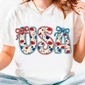 May include: White t-shirt with a red, white, and blue floral design spelling out "USA".