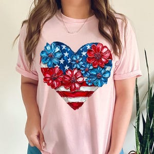 May include: A pink t-shirt with a heart-shaped design featuring a stylized American flag with red, white, and blue flowers.