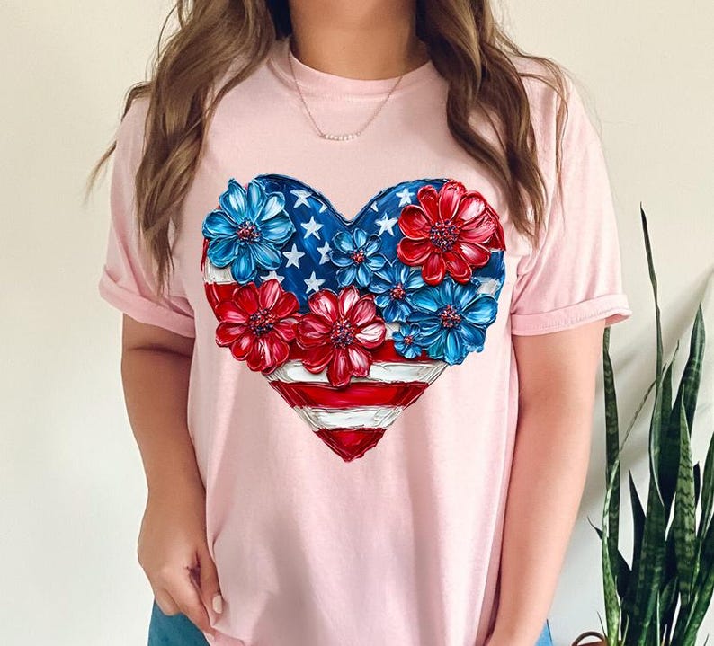 May include: A pink t-shirt with a heart-shaped design featuring a stylized American flag with red, white, and blue flowers.