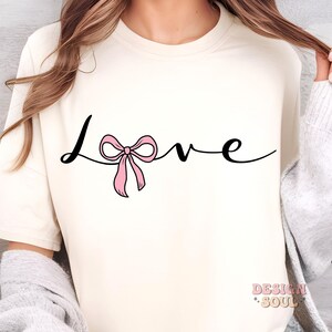 May include: A white t-shirt with the word "love" written in black cursive script. A pink bow is tied around the "o" in the word "love".