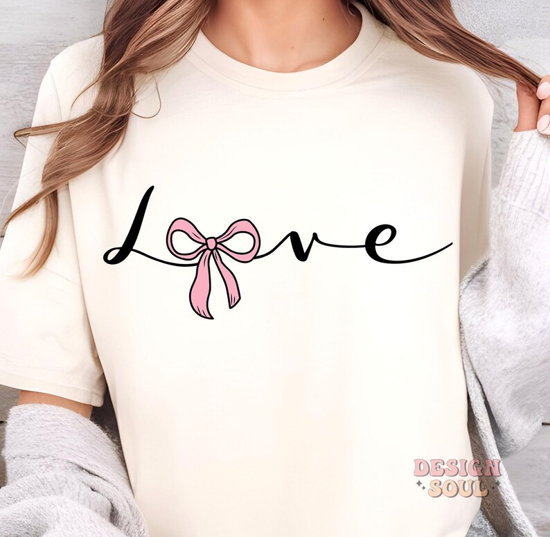 May include: A white t-shirt with the word "love" written in black cursive script. A pink bow is tied around the "o" in the word "love".