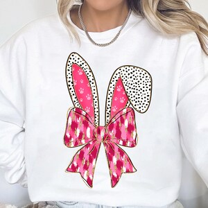 May include: White sweatshirt featuring a pink and gold bunny ear design. The ears are adorned with black polka dots and pink paw prints.