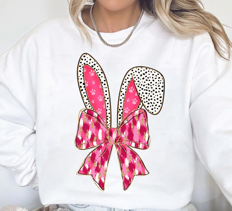May include: White sweatshirt featuring a pink and gold bunny ear design. The ears are adorned with black polka dots and pink paw prints.