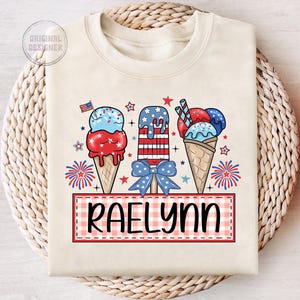 May include: A white t-shirt with a red, white, and blue ice cream cone design. The ice cream cones are decorated with stars and stripes. The text "RAELYN" is written in black on a red and white checkered background.