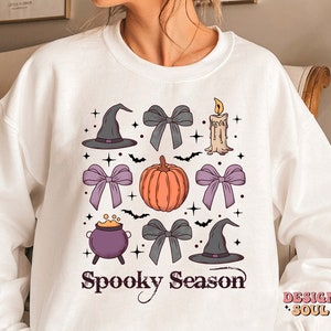 Puede incluir: Sudadera blanca con un dise&ntilde;o de temporada de miedo con una calabaza, sombreros de bruja, lazos, un caldero y una vela. El texto "Spooky Season" est&aacute; impreso en negro.