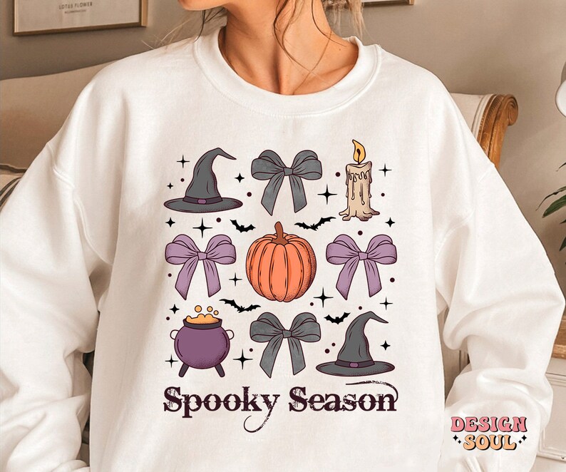 Puede incluir: Sudadera blanca con un dise&ntilde;o de temporada de miedo con una calabaza, sombreros de bruja, lazos, un caldero y una vela. El texto "Spooky Season" est&aacute; impreso en negro.