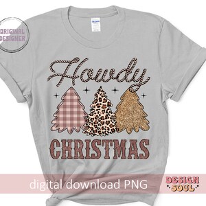 May include: A gray t-shirt with the text "Howdy Christmas" in a brown rope-like font. Three Christmas trees are depicted in the center of the shirt. The trees are pink and white plaid, leopard print, and gold glitter.