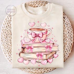 K&ouml;nnte beinhalten: Ein beiger Pullover mit einer rosa und wei&szlig;en Illustration einer Tasse Kaffee, einem Stapel B&uuml;cher und Herzen.