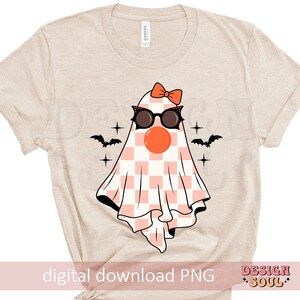 May include: A beige t-shirt with a pink and white checkered ghost wearing sunglasses and a pink bow. The ghost has a red nose and is surrounded by stars and bats.