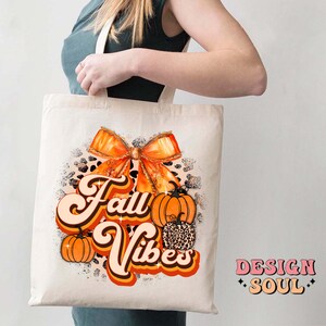 May include: A white tote bag with an orange and brown bow, pumpkins, and the text "Fall Vibes" in a retro font. The bag is made of canvas and has a brown leather handle.