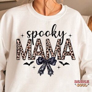 May include: White sweatshirt with the text "spooky mama" in black letters with a leopard print pattern. The text is surrounded by black stars and a black bow with white lace. There are also two black bats on either side of the bow.
