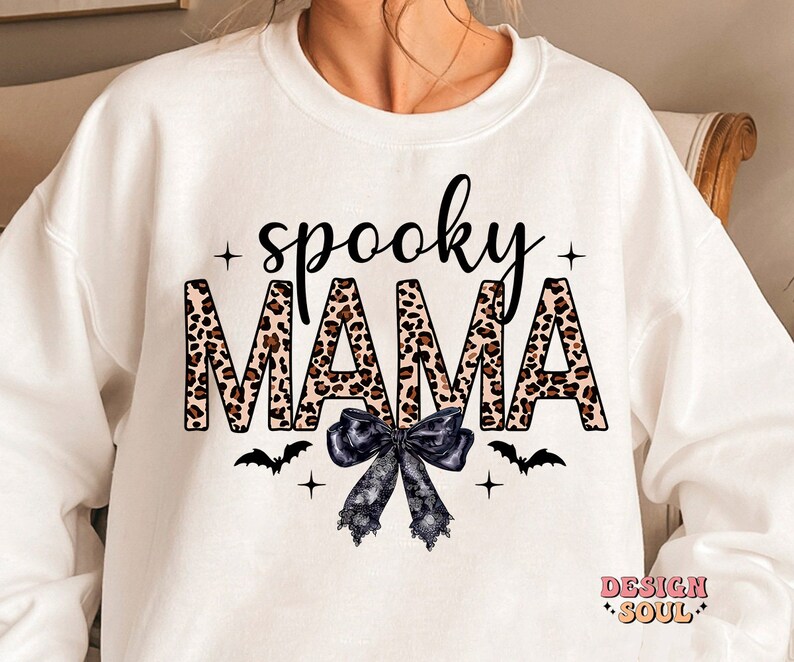 May include: White sweatshirt with the text "spooky mama" in black letters with a leopard print pattern. The text is surrounded by black stars and a black bow with white lace. There are also two black bats on either side of the bow.