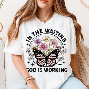 May include: White t-shirt with a graphic design featuring a butterfly with pink and white wings surrounded by flowers. The text "In the waiting God is working" is printed in black on the shirt.