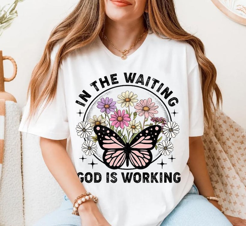 May include: White t-shirt with a graphic design featuring a butterfly with pink and white wings surrounded by flowers. The text "In the waiting God is working" is printed in black on the shirt.