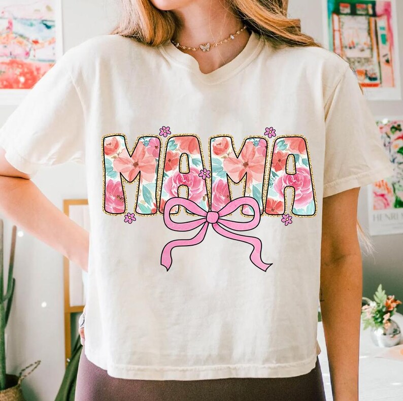 May include: A white t-shirt with a floral print design that says "MAMA" with a pink bow underneath.