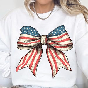 Peut inclure: Sweat-shirt blanc avec un graphique de n&oelig;ud rouge, blanc et bleu. Le n&oelig;ud est dessin&eacute; dans un style vintage et ressemble au drapeau am&eacute;ricain.