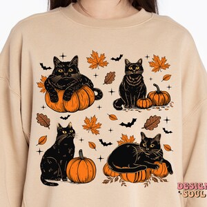 May include: Beige sweatshirt featuring a Halloween-themed design. The graphic includes four black cats, orange pumpkins, autumn leaves, and bats. The design has a vintage, cartoon-like style, perfect for fall.